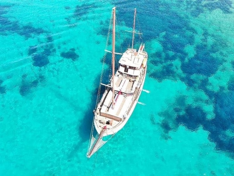A two-masted wooden boat at anchor over pale sand and dark patches of seagrass, seen from above